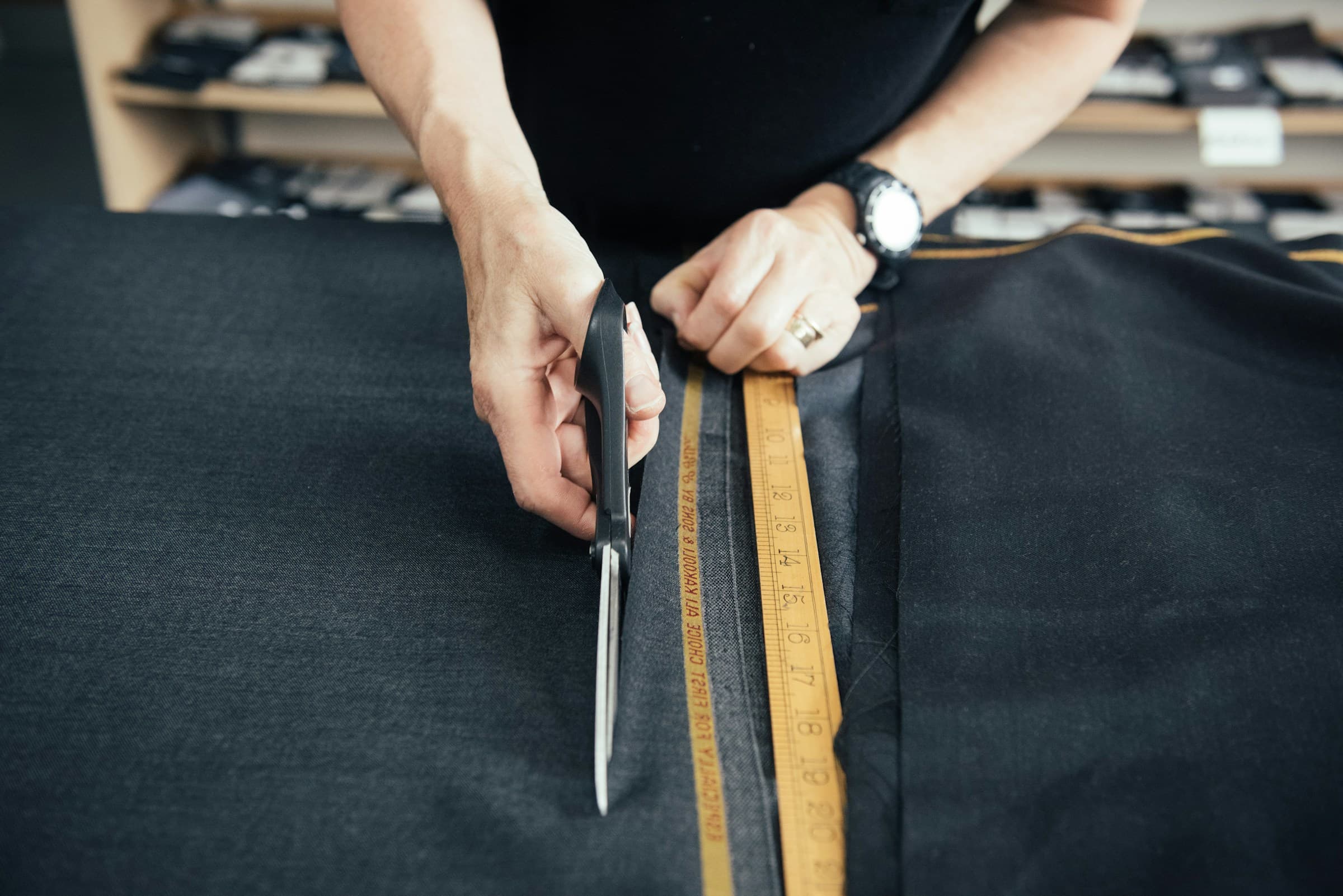 Hands cutting dark fabric with tailor’s shears along a ruler.